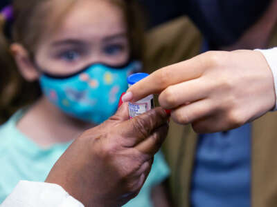 A girl in a mask watches an adult hand over a vaccine vial to another adult