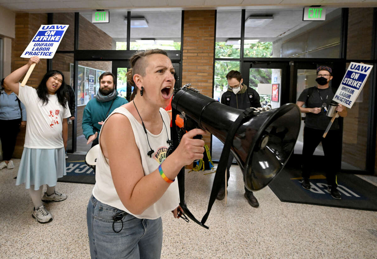 A protester chants into a megaphone during an indoor protest, as others stand behind them with signs reading "UAW ON STRIKE: UNFAIR LABOR PRACTICES"