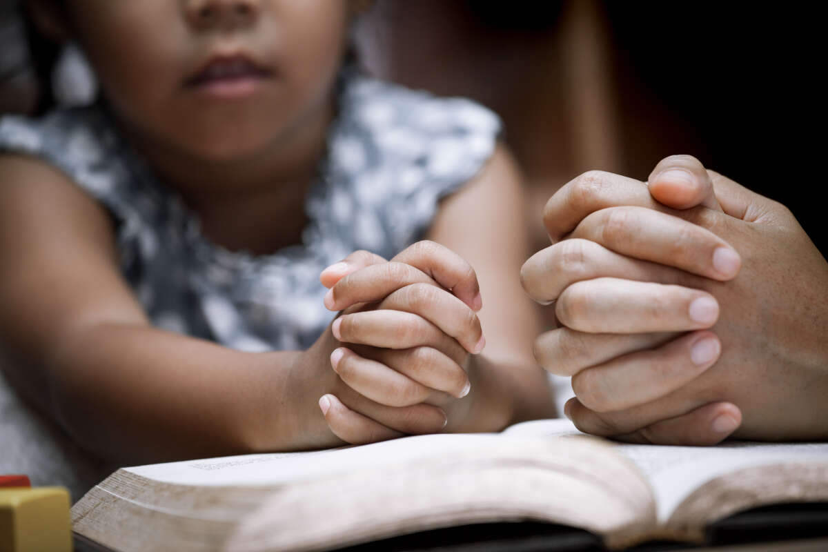 Child and parent hold hands in prayer over bible