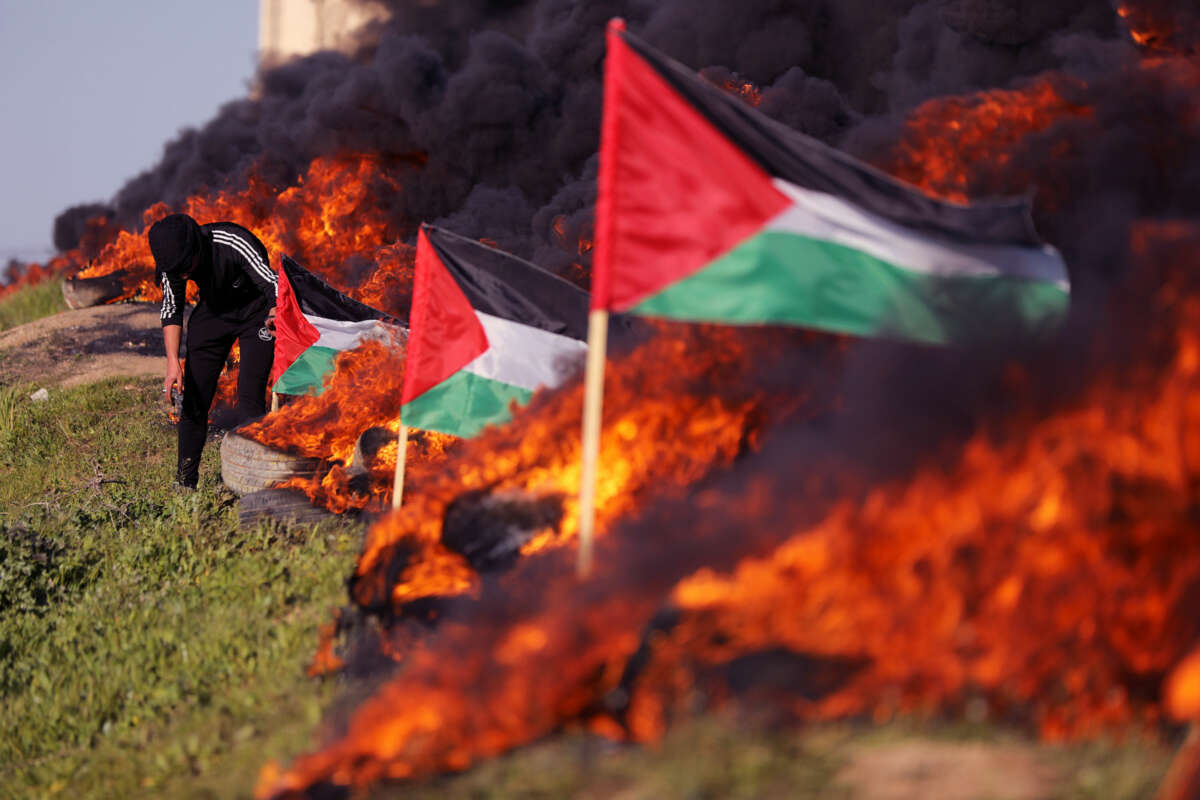A man in a black hoodie kneels amidst Palestinian flags and the smoke tires burning nearby