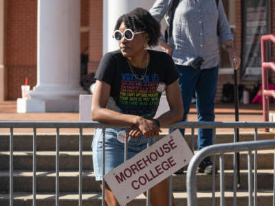 A woman leans against a fence, behind a dangling sign reading "MOREHOUSE COLLEGE"