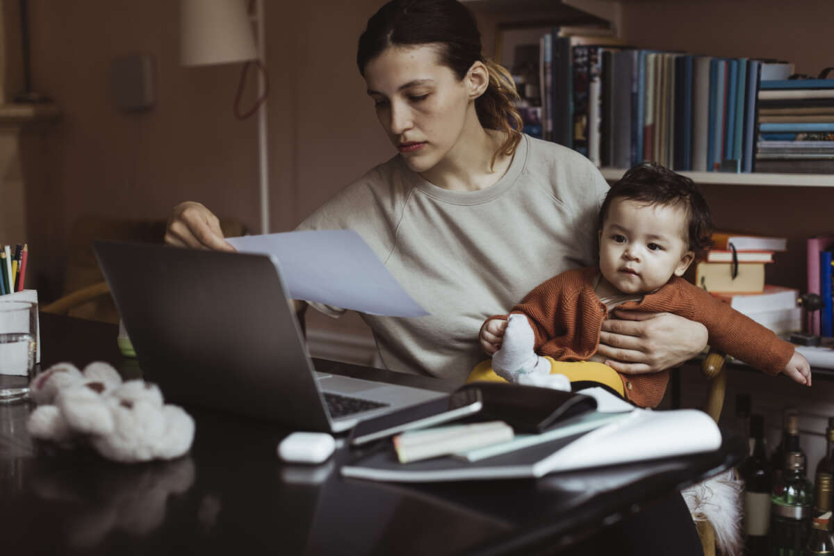 An overworked mom sits at a table with a pile of papers holding a baby