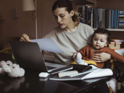 An overworked mom sits at a table with a pile of papers holding a baby