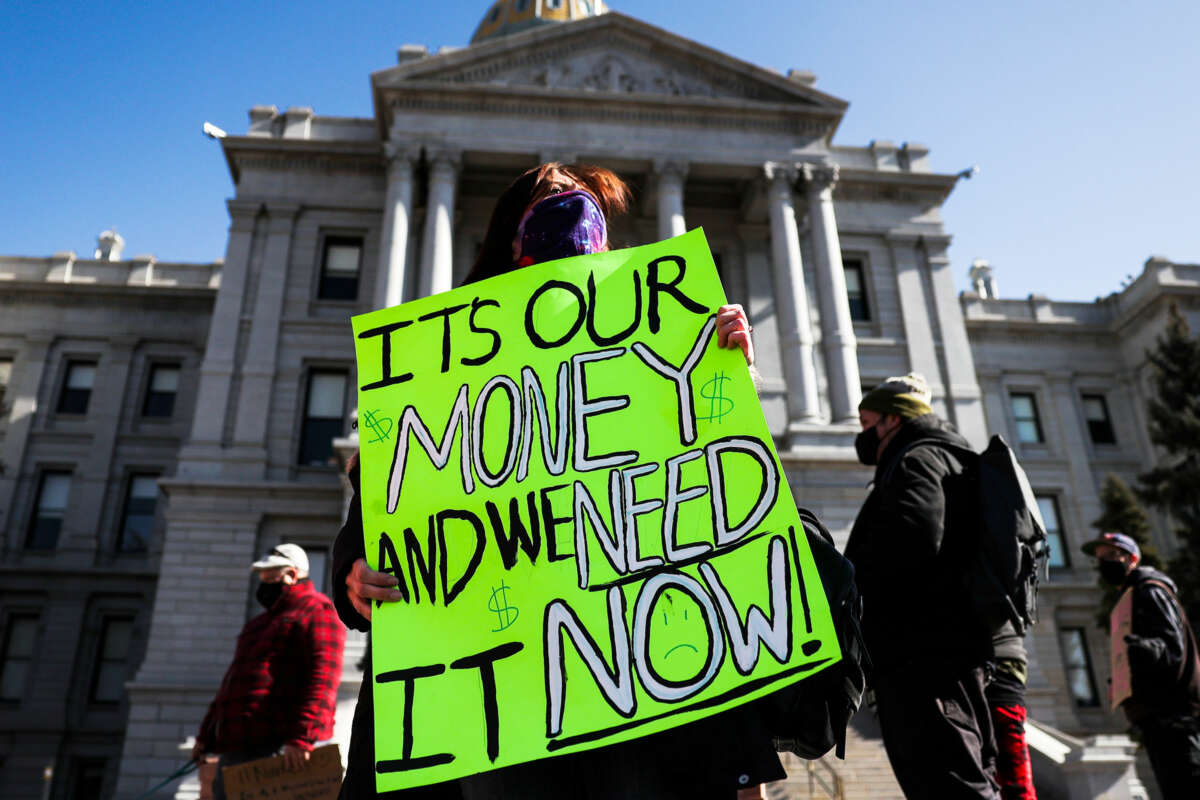 A masked woman on the steps of a state capitol holds a green sign reading "IT'S OUR MONEY AND WE WANT IT NOW" during an outdoor protest