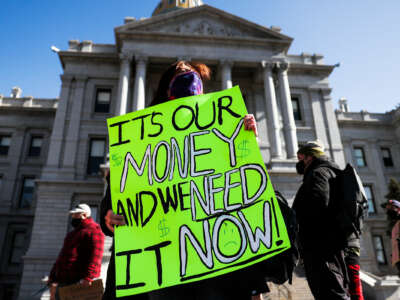 A masked woman on the steps of a state capitol holds a green sign reading "IT'S OUR MONEY AND WE WANT IT NOW" during an outdoor protest