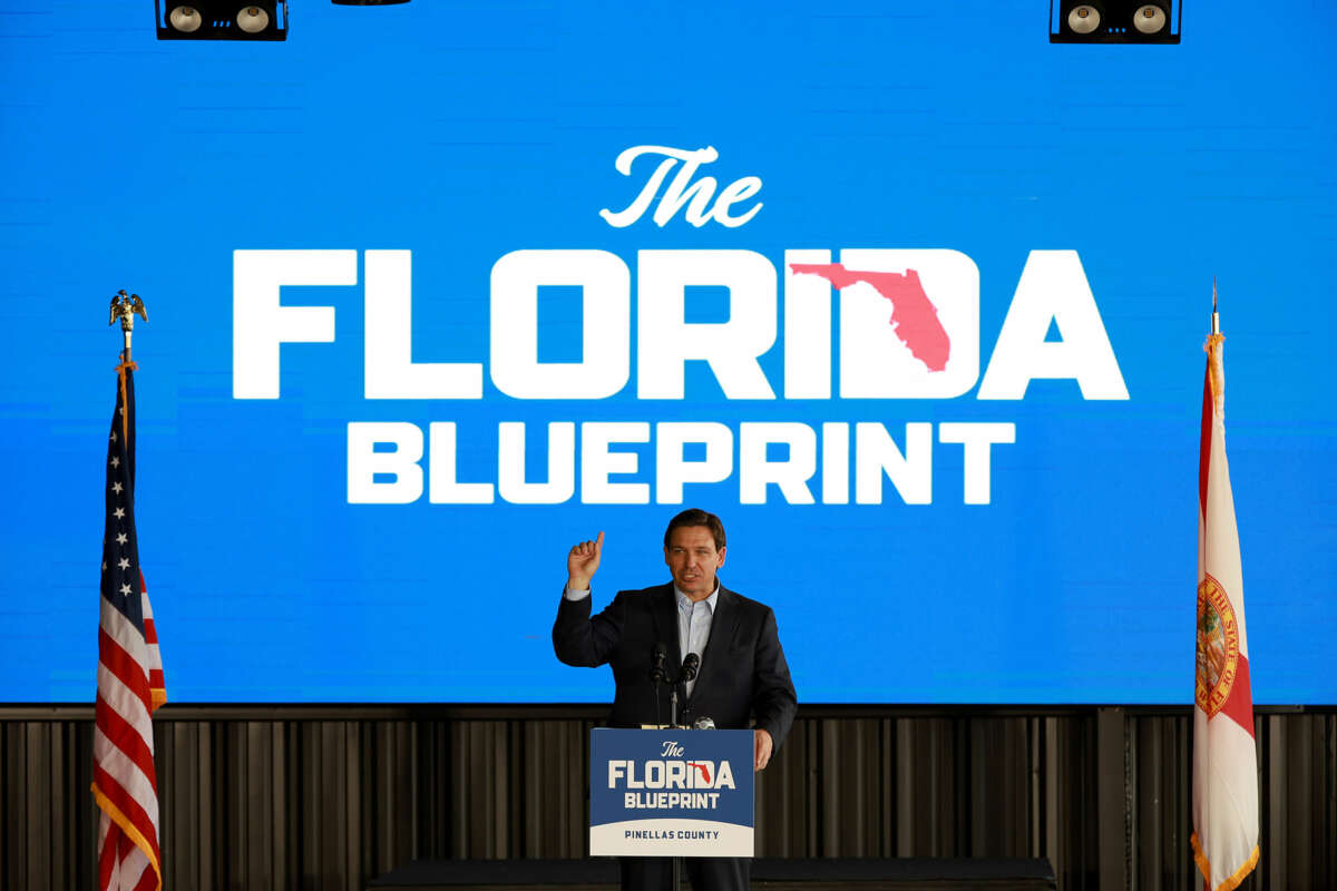 Ron Desantis stands in front of a sign reading "THE FLORIDA BLUEPRINT" while he speaks to an audience