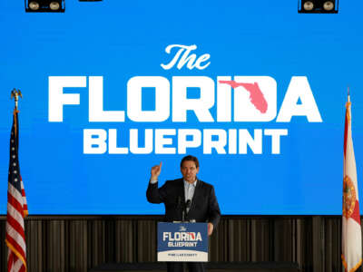 Ron Desantis stands in front of a sign reading "THE FLORIDA BLUEPRINT" while he speaks to an audience