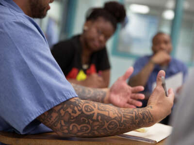 The tattooed arm of a man holding a pen is seen as he explains something to others seated at a table with him