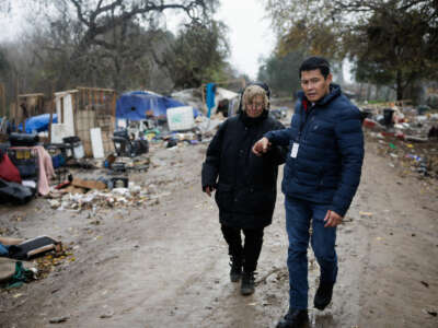 A man leads a woman by the hand through a homeless encampment in rainy weather
