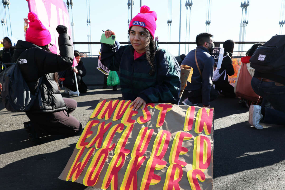 A person in a pink hat displays a sign reading "INVEST IN EXCLUDED WORKERS" while kneeling during an outdoor protest