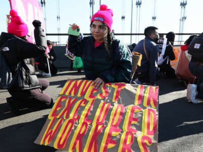 A person in a pink hat displays a sign reading "INVEST IN EXCLUDED WORKERS" while kneeling during an outdoor protest