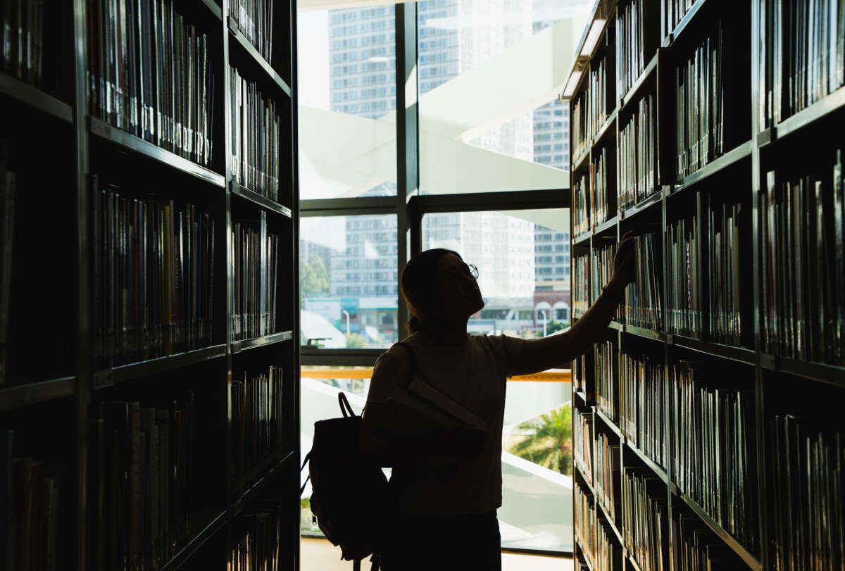 A person looks at books on shelves in a library, silhouetted by light from window behind them