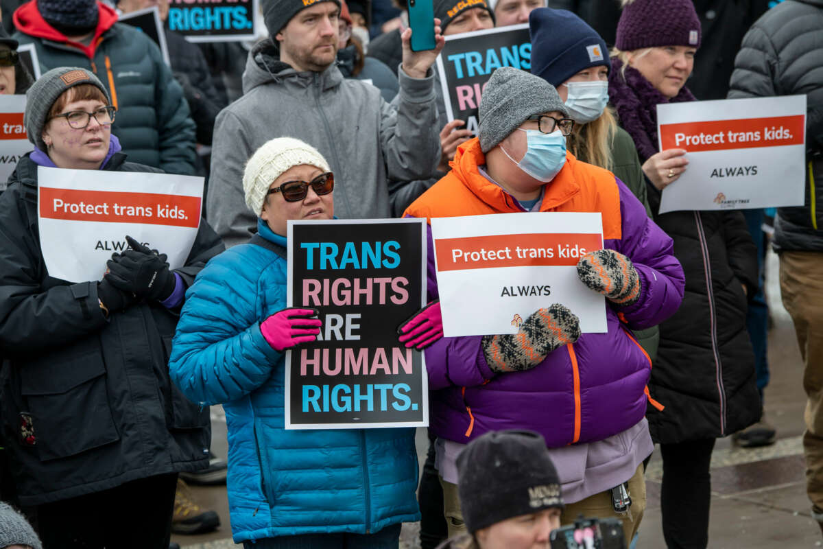 People in winter wear display signs reading "TRANS RIGHTS ARE HUMAN RIGHTS" and "PROTECT TRANS KIDS ALWAYS" during an outdoor protest