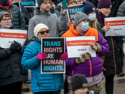 People in winter wear display signs reading "TRANS RIGHTS ARE HUMAN RIGHTS" and "PROTECT TRANS KIDS ALWAYS" during an outdoor protest