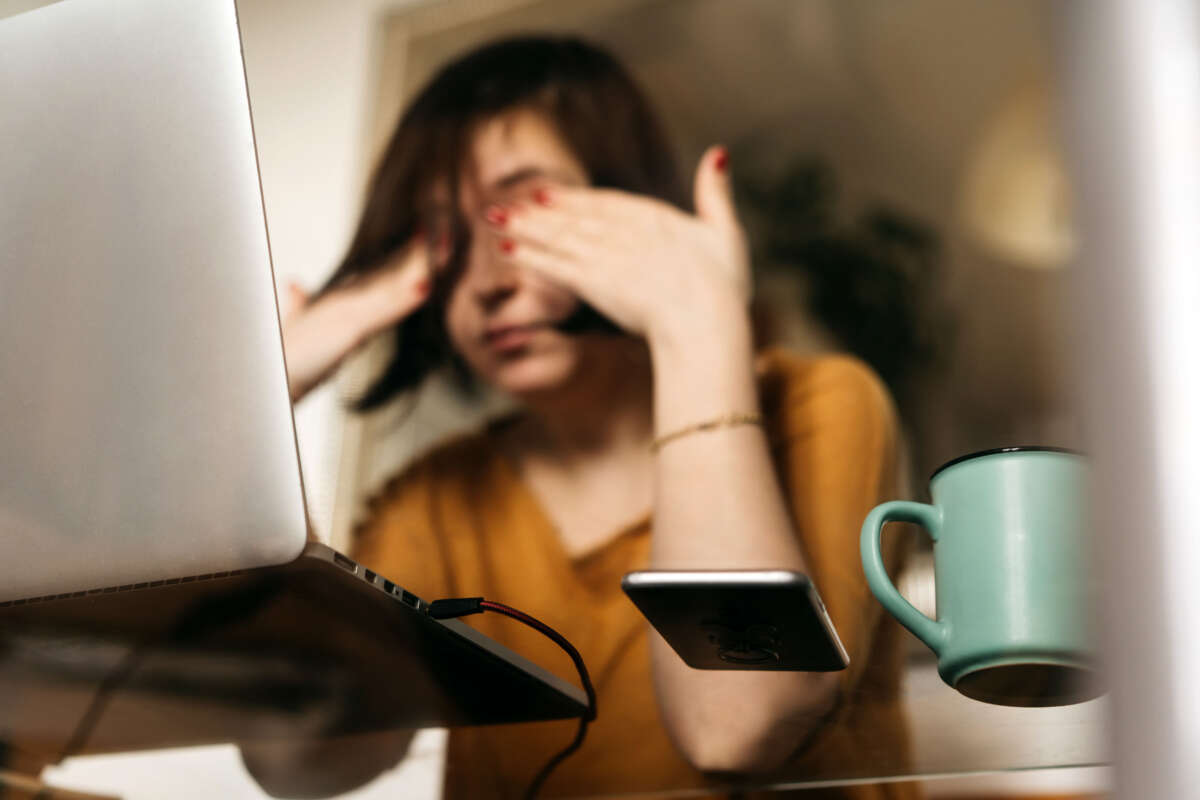 Low angle of woman covering her eyes while seated at glass desk facing laptop computer. A phone and coffee mug sit by the computer.