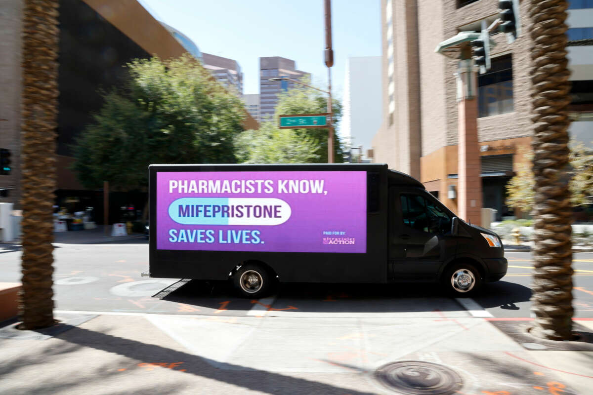 A truck with a video screen reading "PHARMACISTS KNOW, MIFERPRISTONE SAVES LIVES" drives through a city center