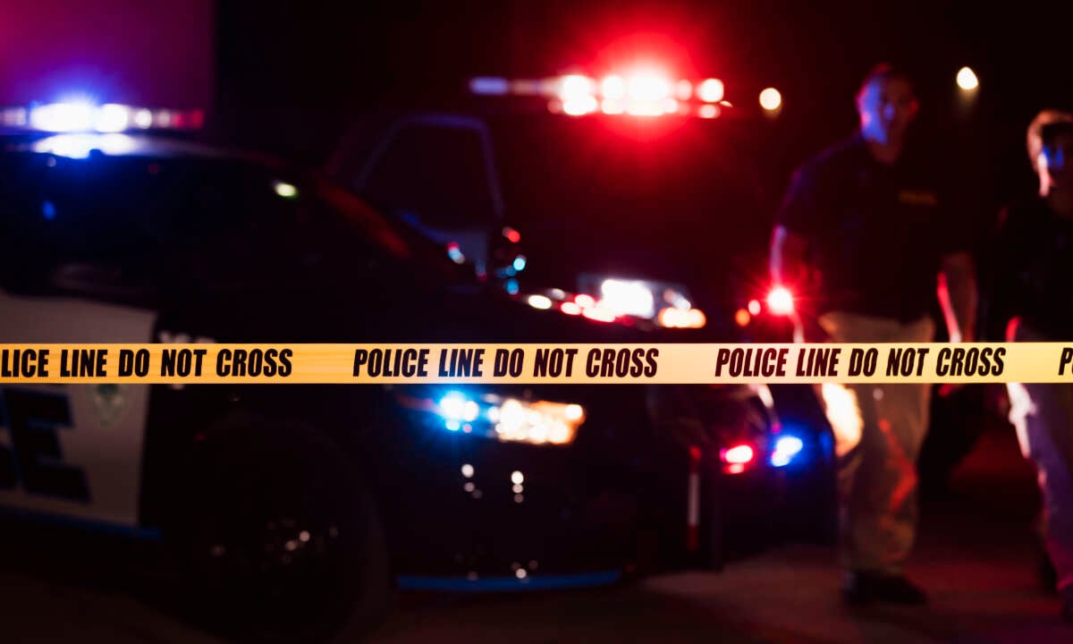 Two police officers standing in front of patrol cars, behind police tape, wearing bulletproof vests and duty belts. It is nighttime and the emergency lights on top of the vehicles are flashing.