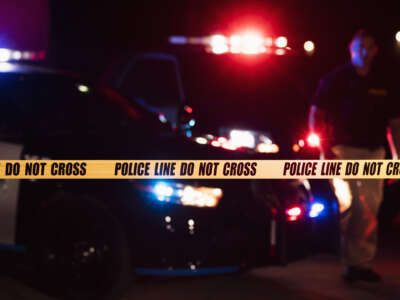 Two police officers standing in front of patrol cars, behind police tape, wearing bulletproof vests and duty belts. It is nighttime and the emergency lights on top of the vehicles are flashing.