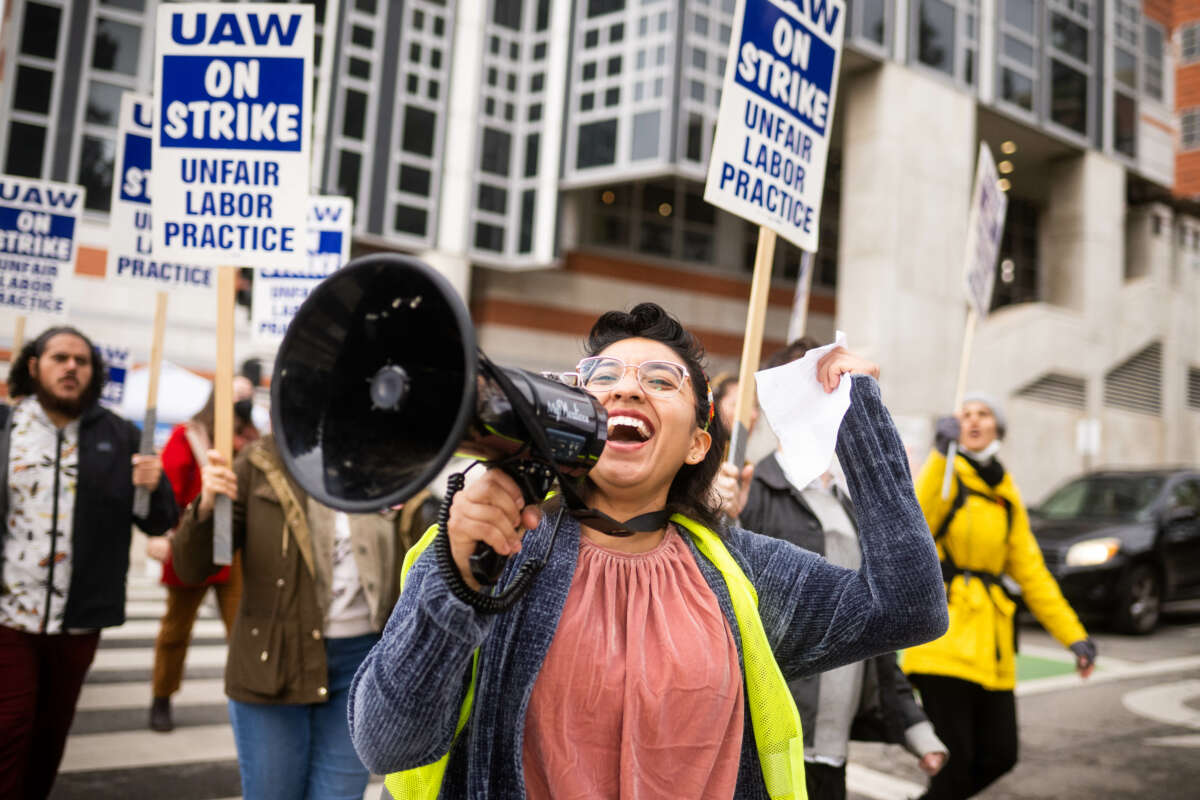 An activist hold a bullhorn and raises a fist in the air in a crowd of people holding signs that read "UAW on strike"
