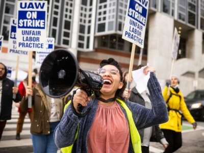 An activist hold a bullhorn and raises a fist in the air in a crowd of people holding signs that read "UAW on strike"