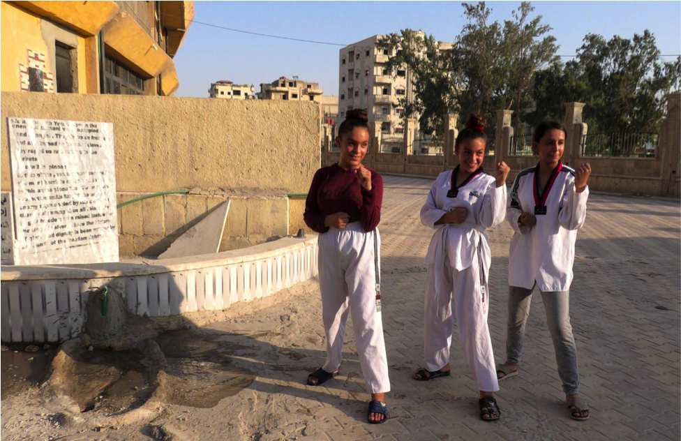 נערות כורדיות וערביות לומדות קראטה באצטדיון של רקה, ששימש בעבר את דאעש (נקרא גם דאעש) כ"כלא מוות". משמאלם אנדרטה לנשים יזידיות המשועבדות לדאעש.
