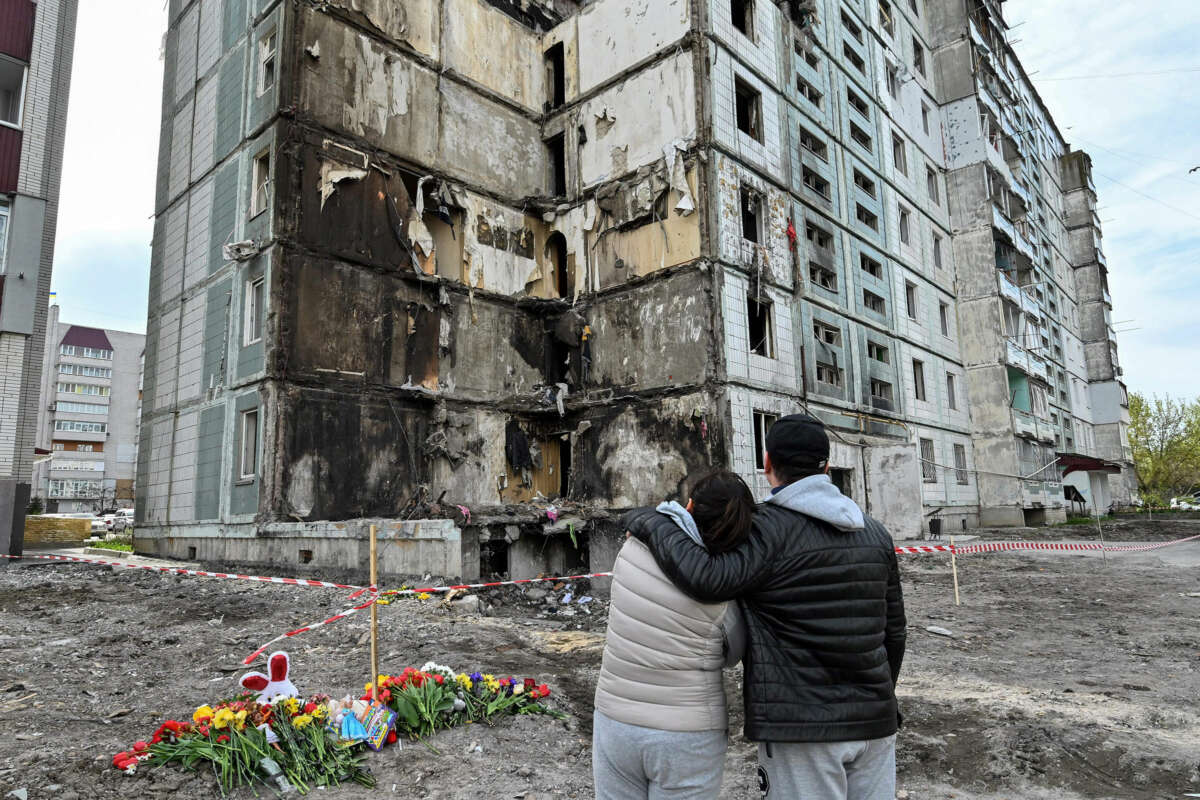 Dos personas se abrazan mientras miran un complejo de apartamentos parcialmente destruido contra un cielo nublado