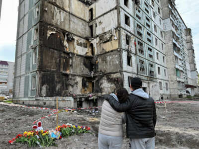 Two people embrace while looking at a partially-destroyed apartment complex against a cloudy sky