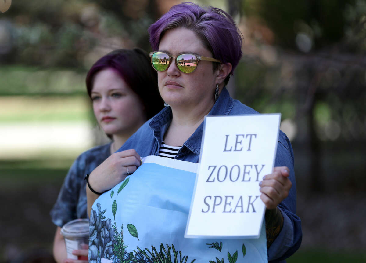 A person holds a sign reading "LET ZOOEY SPEAK" during an outdoor protest