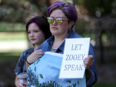 A person holds a sign reading "LET ZOOEY SPEAK" during an outdoor protest