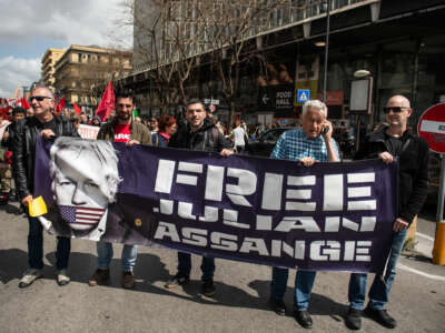 People march behind a banner displaying the face of Julian Assange alongside the words "FREE JULIAN ASSANGE" during an outdoor action