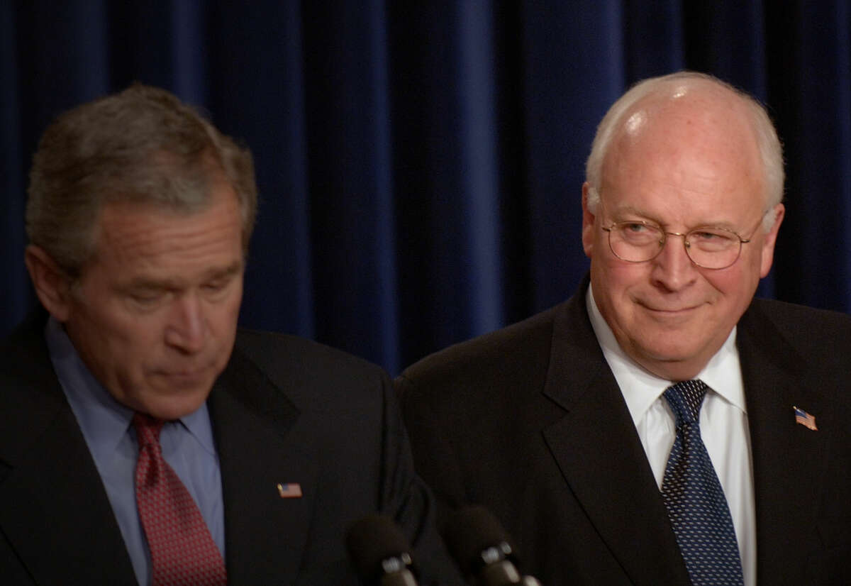 Dick Cheney smirks at George W. Bush, who is reading off of a piece of paper while standing at a podium to deliver a speech