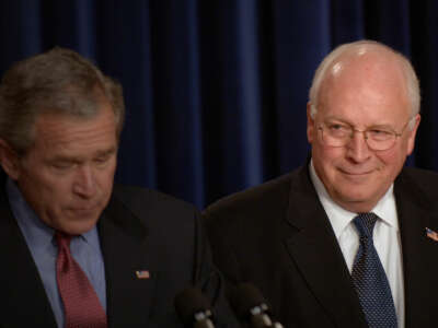 Dick Cheney smirks at George W. Bush, who is reading off of a piece of paper while standing at a podium to deliver a speech