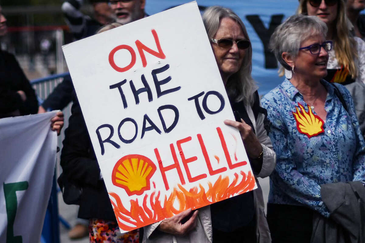 protesters gather as one holds a sign baring the Shell Oil logo and text reading "ON THE ROAD TO HELL"