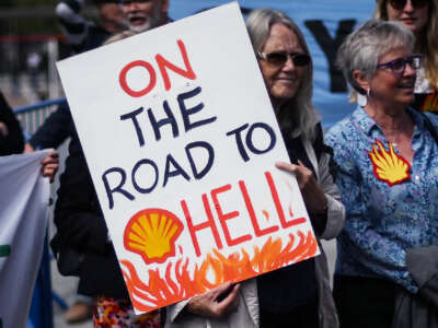 protesters gather as one holds a sign baring the Shell Oil logo and text reading "ON THE ROAD TO HELL"