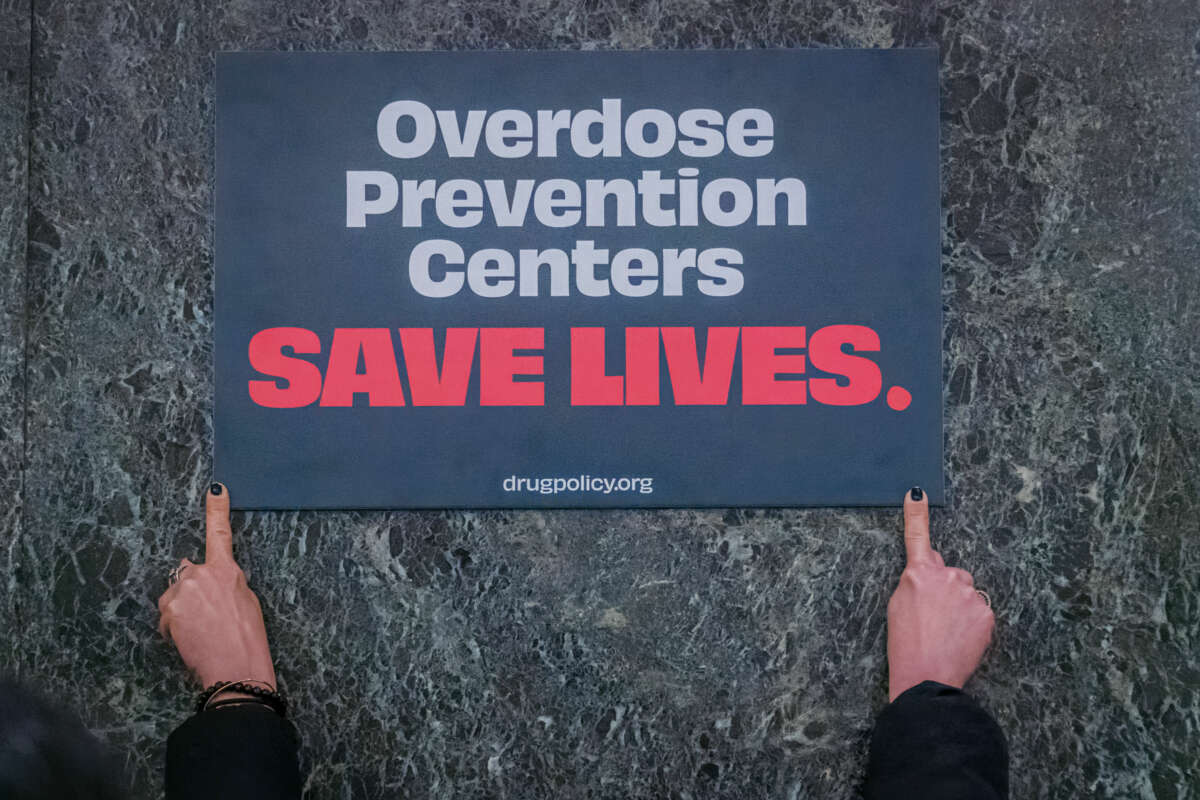 A sign reading "Overdose prevention centers SAVE LIVES." is displayed against a green marble background