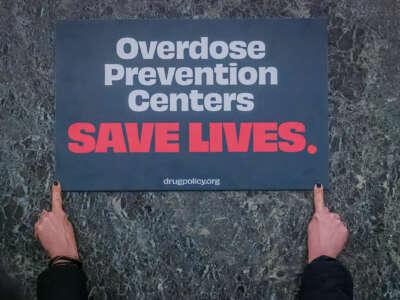 A sign reading "Overdose prevention centers SAVE LIVES." is displayed against a green marble background