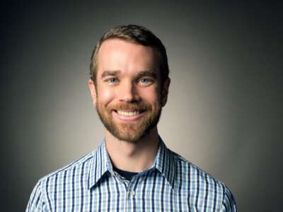 a head shot of a white person with dark blonde short hair and a beard smiles against a vignette background.