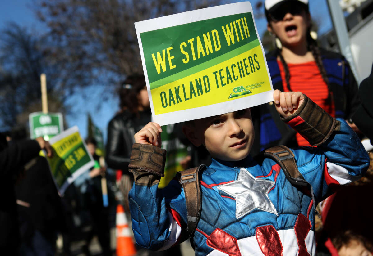 A little boy wearing a Captain America costume holds a sign at a protest saying "We stand with Oakland teachers"
