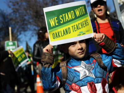 A little boy wearing a Captain America costume holds a sign at a protest saying "We stand with Oakland teachers"