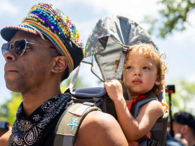 A parent wearing a rainbow hat carries their child on their back at a protest for LGBTQ rights