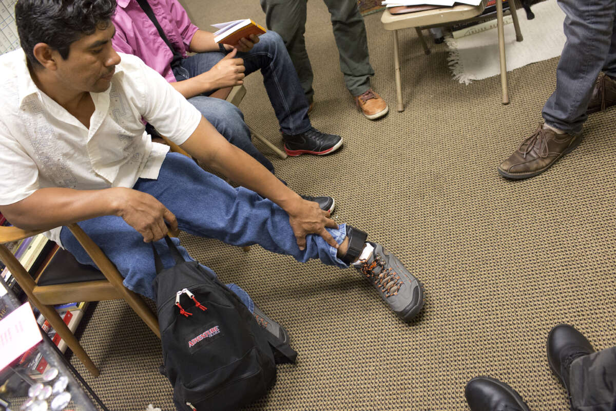 A man sits with a backpack, reaching to touch his ankle monitor.