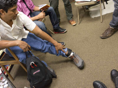 A man sits with a backpack, reaching to touch his ankle monitor.