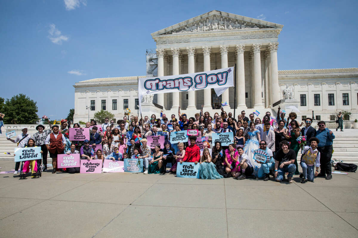 A diverse group of colorfully-dressed people unite under a banner of "TRANS JOY" that they hold above their heads on the steps of the U.S. supreme court building