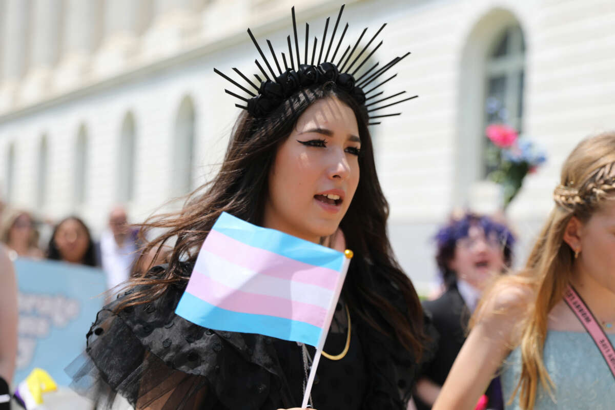 A girl in a black dress with a black flower crown holds the trans flag and marches alongside other trans teens