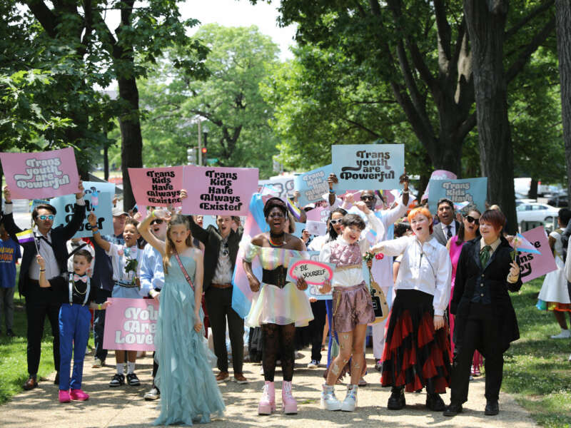 Trans youth, wearing colorful clothing and joyful expressions, march to the capitol holding signs reading "TRANS KIDS HAVE ALWAYS EXISTED" and "TRANS YOUTH ARE POWERFUL"