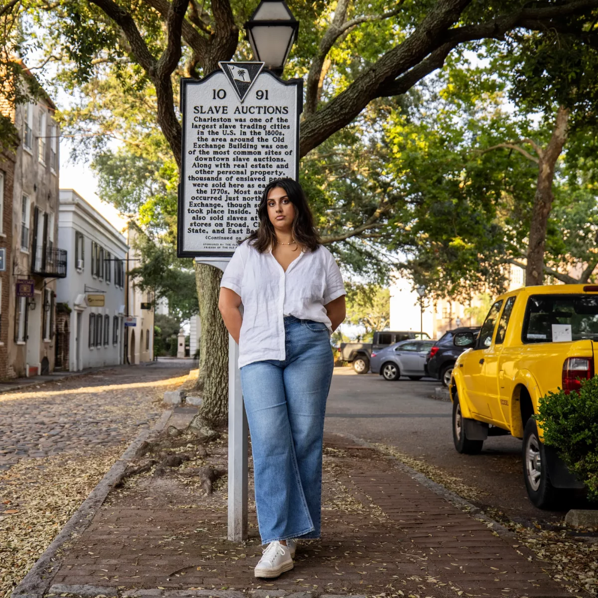 A woman with brown skin stands on a street in Charleston in front of a sign with information about slave auctions