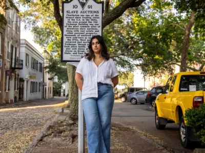 A woman with brown skin stands on a street in Charleston in front of a sign with information about slave auctions