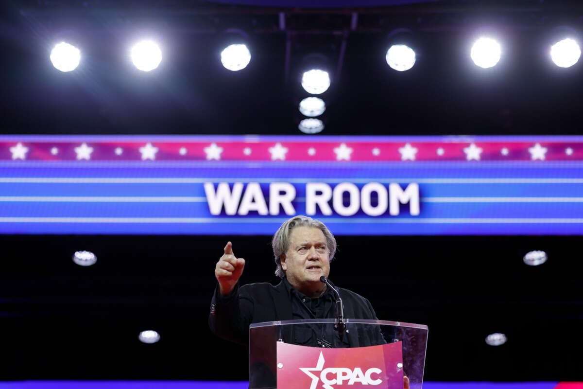 Steve Bannon points out into a crowd as he speaks behind a podium and in front of a lit sign reading "WAR ROOM" during cpac 2023