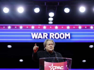 Steve Bannon points out into a crowd as he speaks behind a podium and in front of a lit sign reading "WAR ROOM" during cpac 2023