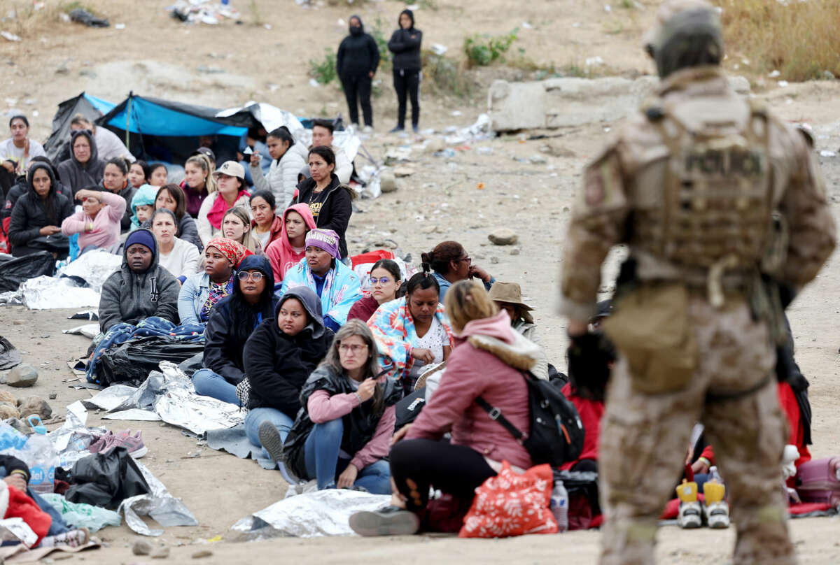 People sit in a line as they're watched by an armed and uniformed border patrol agent